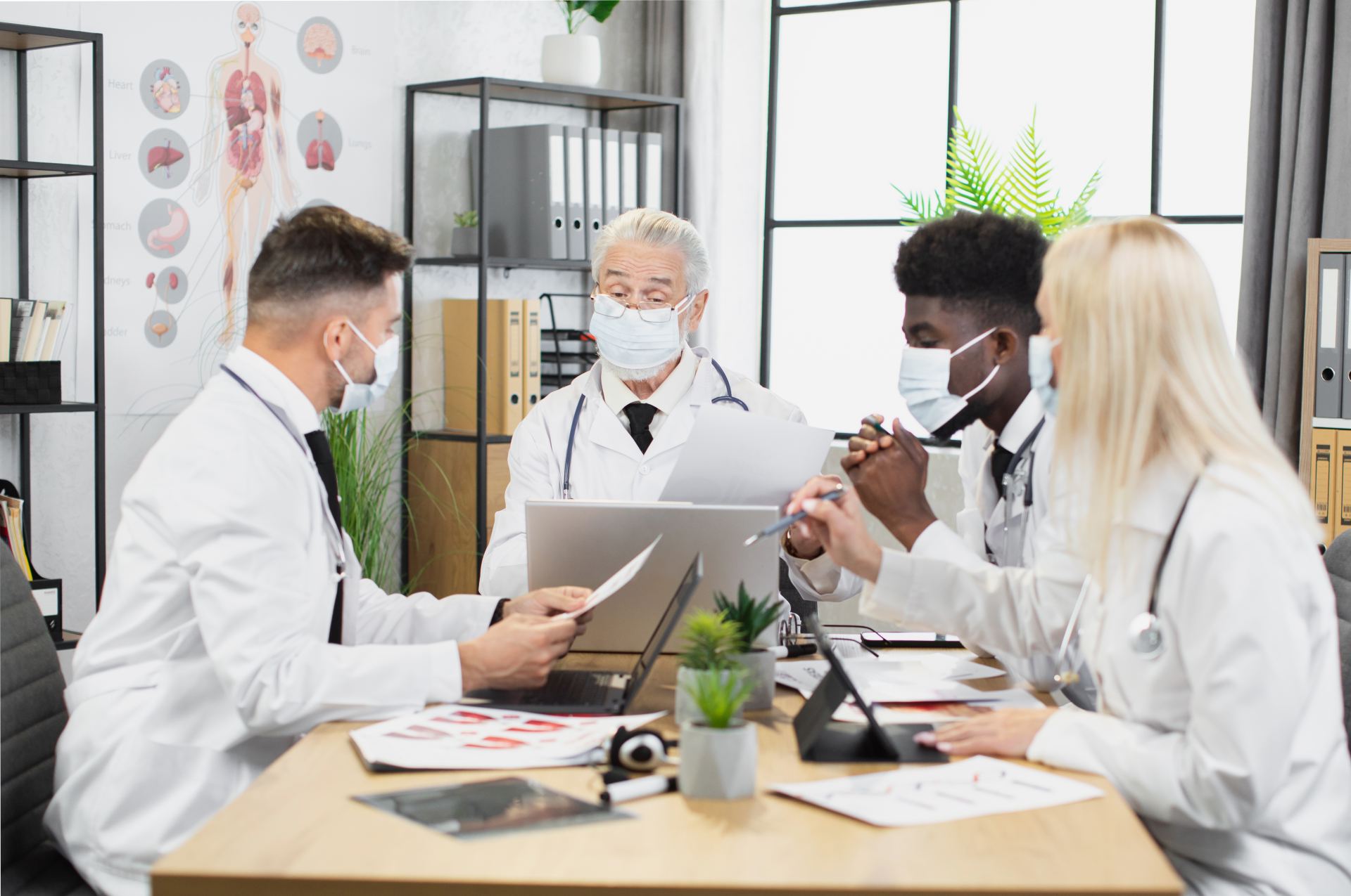 Multiracial healthcare specialists in face mask discussing covid infection while sitting at desk during conference. Male and female doctors using modern gadgets on meeting.